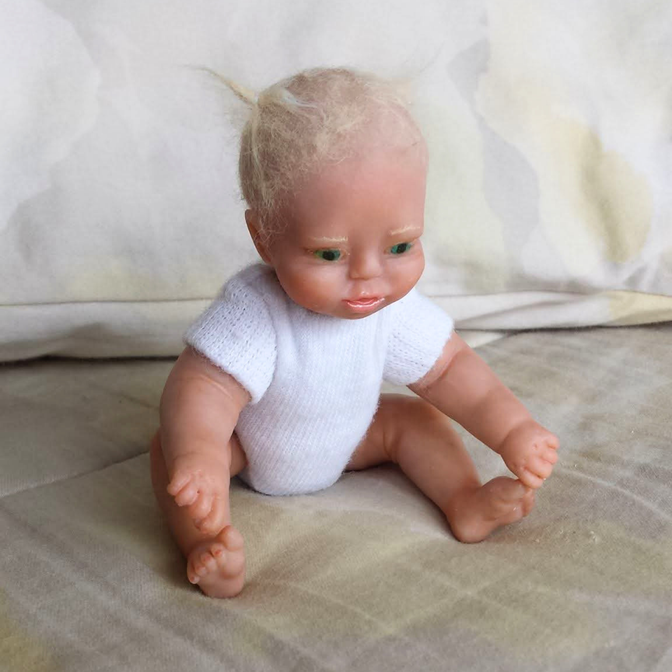 Yana, a six-inch polymer clay toddler doll with blue-green eyes and wispy blond hair, sitting in a white bodysuit.
