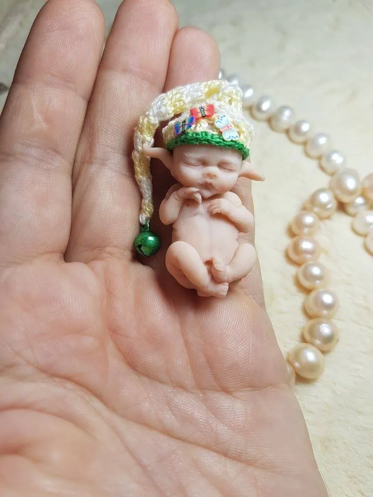Whimsical OOAK miniature baby doll wearing a colorful rainbow-striped crocheted elf hat with green pom pom, held gently in palm with pearl beads - by Mini Baby Dolls