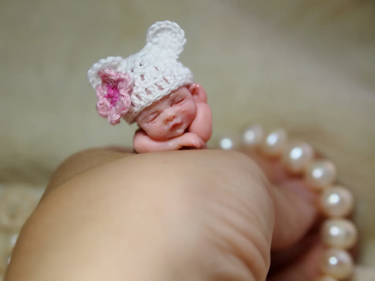 Sleeping OOAK miniature baby doll in white crocheted bear ear hat with pink flower accent, resting on fist with pearl beads - by Mini Baby Dolls