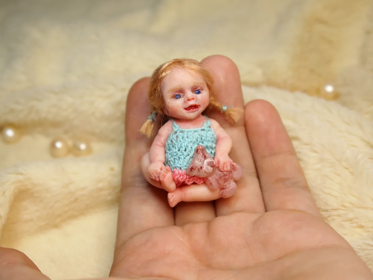 OOAK miniature toddler doll with blonde curly hair and blue eyes, wearing teal crocheted romper, sitting in palm - by Mini Baby Dolls