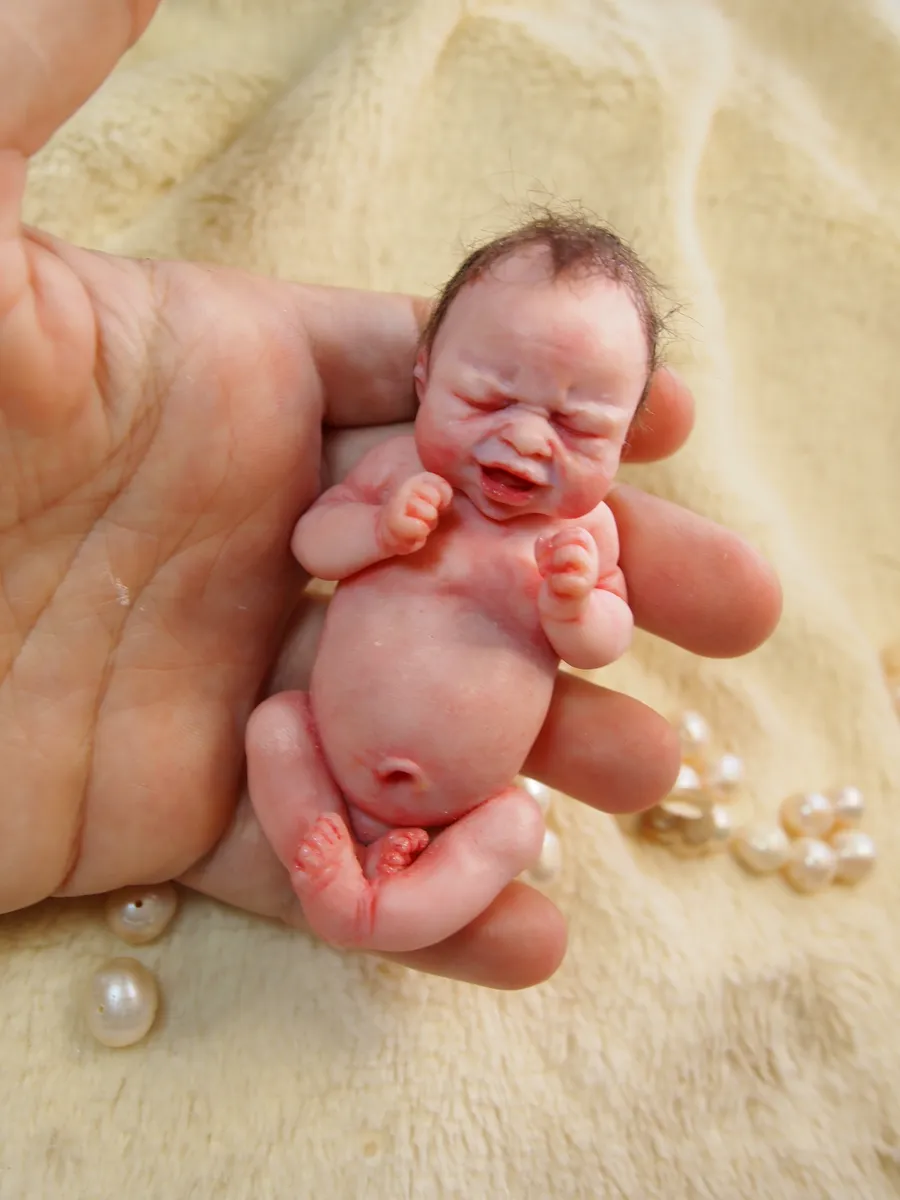 Highly detailed OOAK polymer clay miniature baby doll with realistic crying or laughing expression, held gently in palm with pearl beads, showcasing expressive facial sculpting - by Mini Baby Dolls