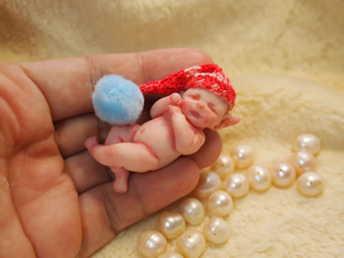 Sleeping OOAK miniature baby doll in red crocheted outfit with blue pom pom accent, held gently in palm - by Mini Baby Dolls