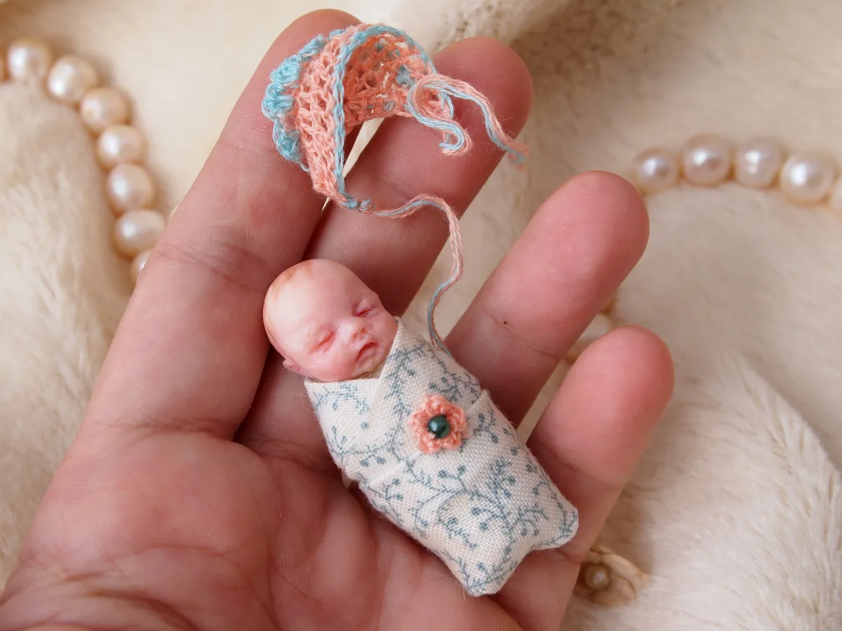 Sleeping OOAK miniature baby doll tucked into a tiny handmade fabric sleeping bag with floral pattern, wearing a peach crocheted bonnet, held gently between fingers - by Mini Baby Dolls