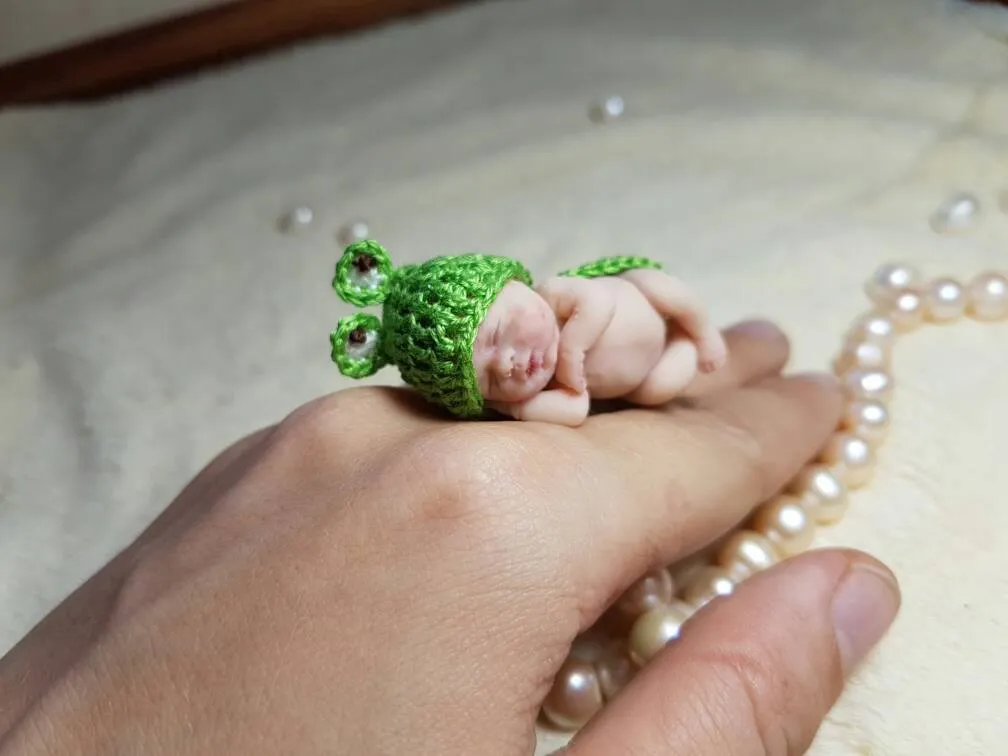 Sleeping OOAK miniature baby doll in adorable green crocheted frog costume with big googly eyes, resting on back of hand with pearl beads - by Mini Baby Dolls