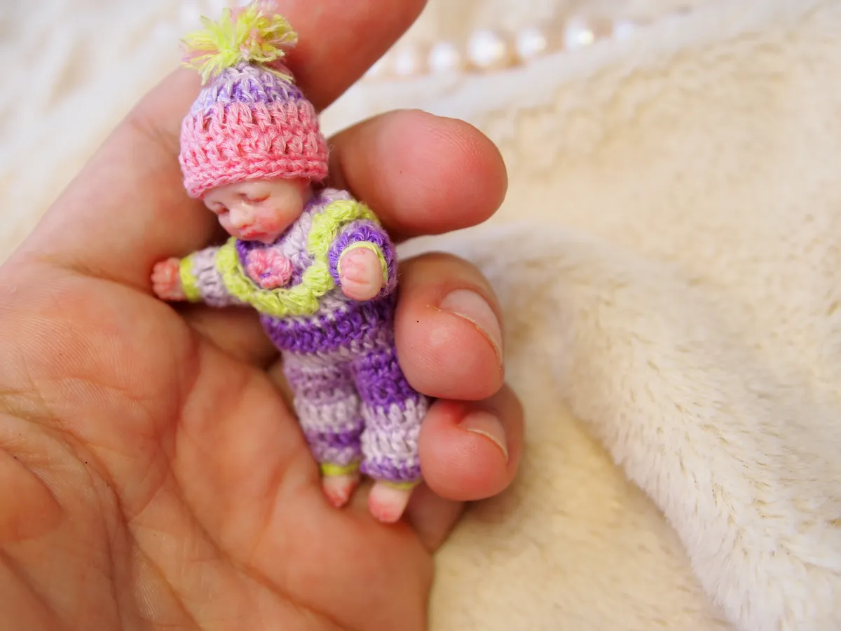 Sleeping OOAK miniature baby doll in pastel pink and purple crocheted outfit with matching pink bonnet, held gently in hand - by Mini Baby Dolls