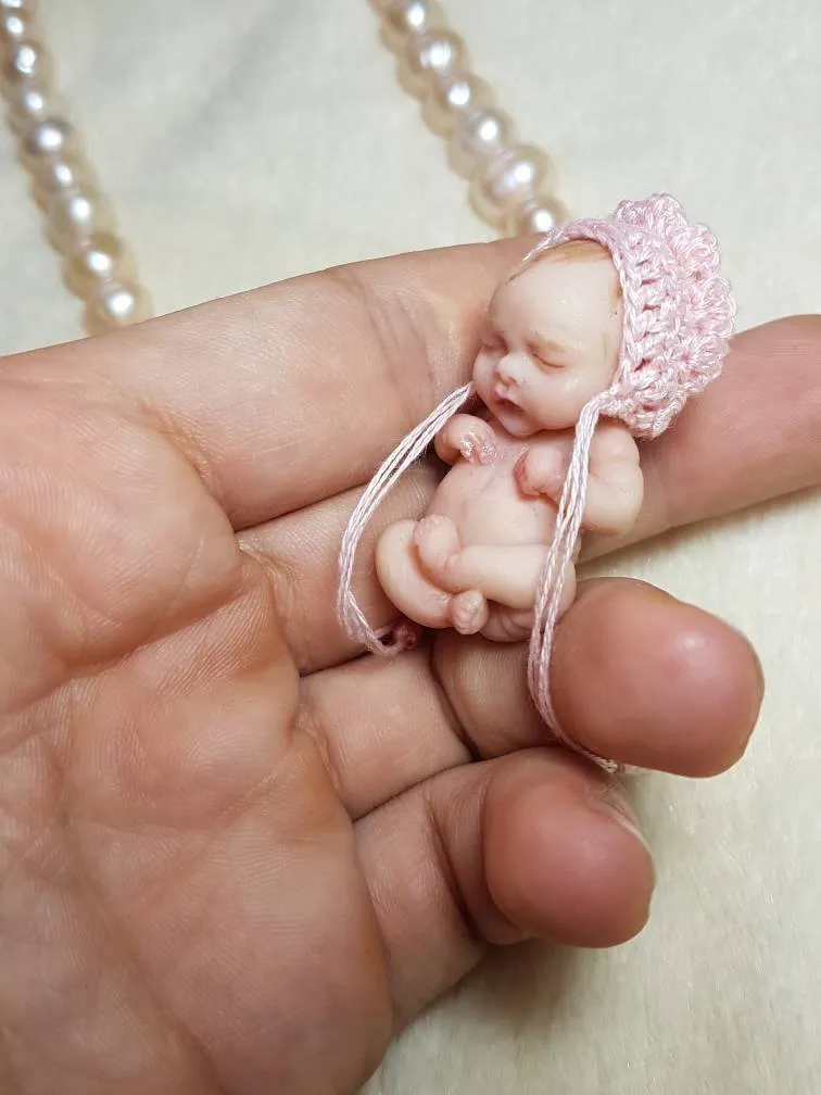 Sleeping OOAK miniature baby doll wearing a delicate pink crocheted bonnet with ribbon ties, displayed in palm with pearl beads - by Mini Baby Dolls