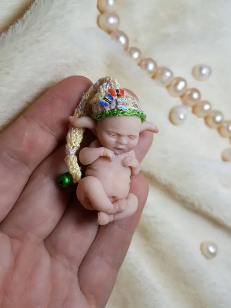 Sleeping OOAK miniature baby doll wearing a colorful handmade crocheted elf hat with rainbow stripes and green pom pom, held gently in palm with pearl beads - by Mini Baby Dolls