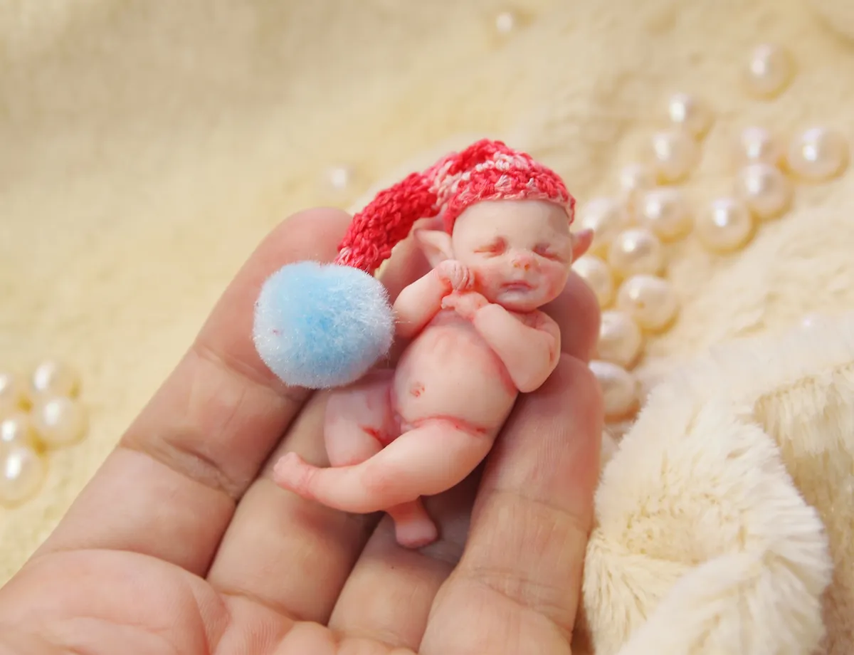 Sleeping OOAK polymer clay miniature baby doll wearing a bright red crocheted hat, cuddling a soft blue pom pom, displayed with pearl beads - by Mini Baby Dolls