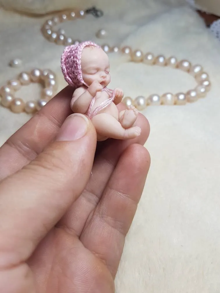 Sleeping OOAK miniature baby doll wearing a sparkly pink crocheted bonnet, held delicately between fingers with pearl beads - by Mini Baby Dolls