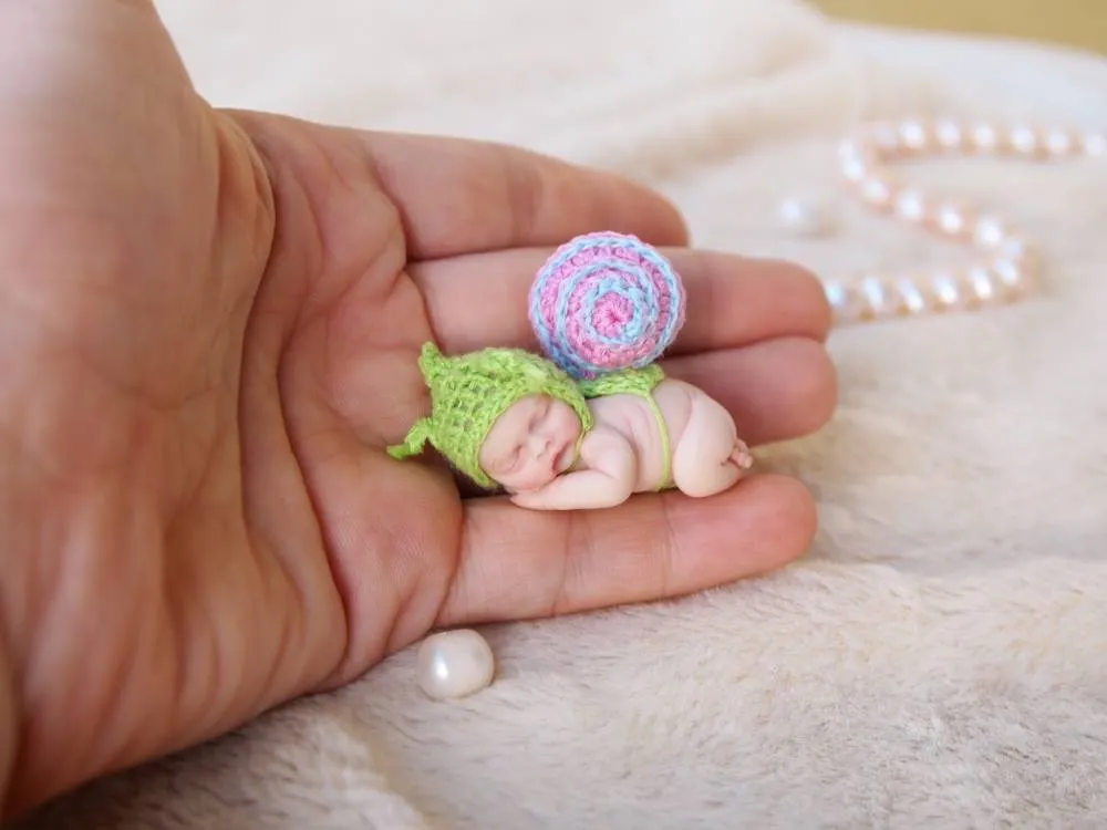 Whimsical OOAK miniature baby doll in green crocheted outfit with colorful striped snail shell on back, held in palm - by Mini Baby Dolls