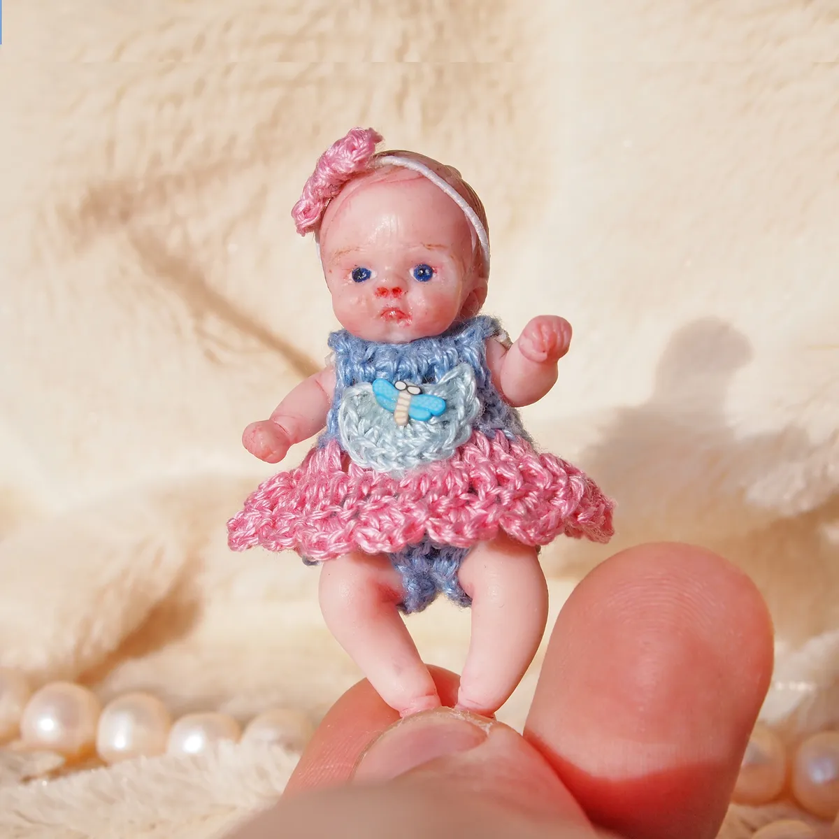 Standing miniature polymer clay baby doll with blue eyes wearing a handmade grey-blue and pink crocheted dress with matching pink bow headband - by Mini Baby Dolls