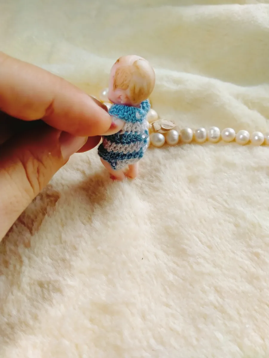 Cheerful OOAK miniature baby doll in blue and grey striped crocheted romper, held between fingers with pearl beads - by Mini Baby Dolls