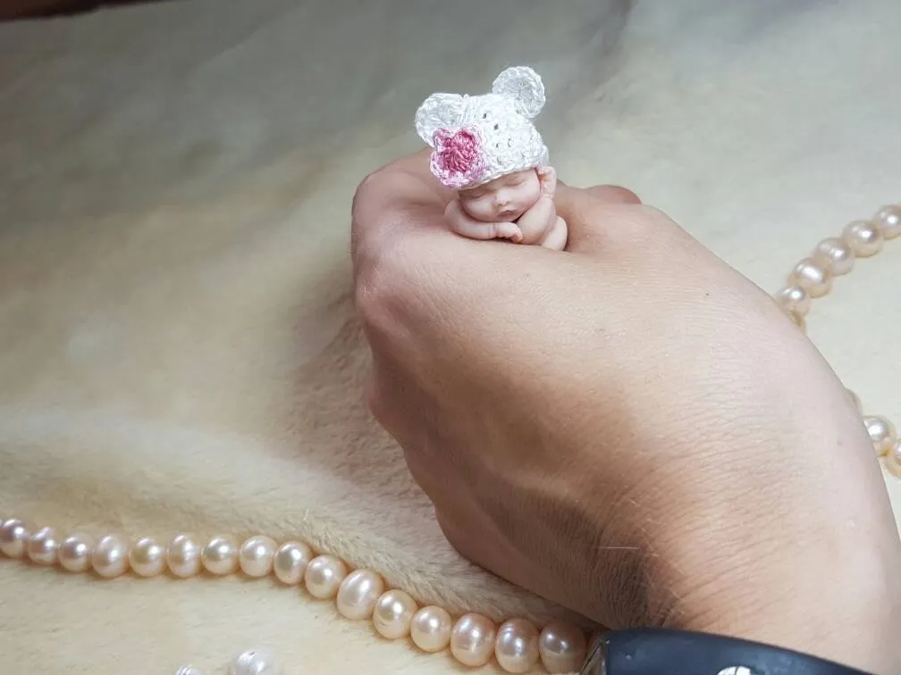 Sweet OOAK miniature baby doll wearing a white crocheted bear-ear hat adorned with a pink flower, resting in palm with pearl beads - by Mini Baby Dolls
