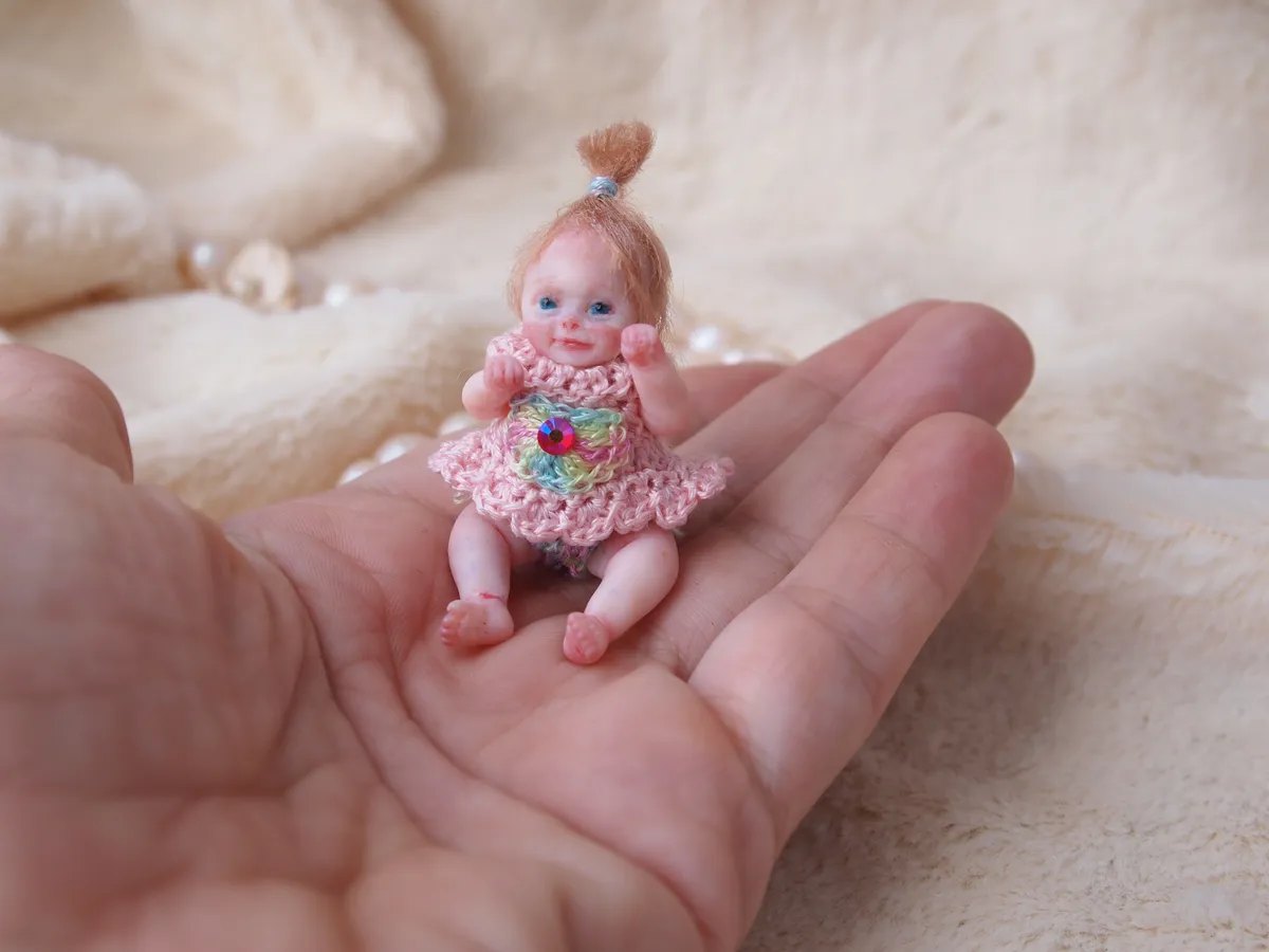 Adorable OOAK miniature toddler doll with auburn ponytail and blue eyes, wearing pink crocheted dress with rainbow flower detail, sitting in palm - by Mini Baby Dolls
