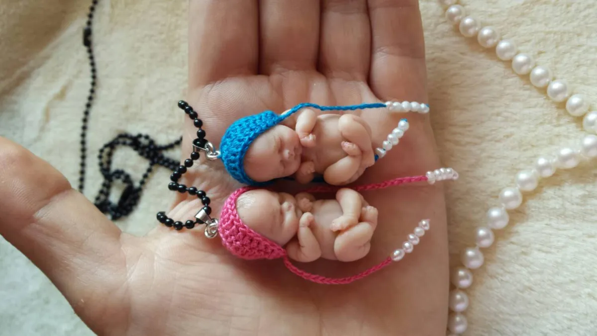Two ultra-tiny OOAK miniature baby dolls in matching crocheted outfits - one in blue, one in pink - displayed together with delicate chain necklace and pearl beads - by Mini Baby Dolls