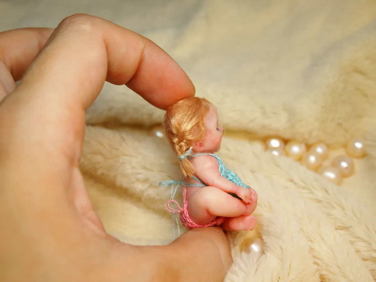 Ultra tiny OOAK miniature blonde baby doll held delicately between fingers, showcasing incredible miniature scale detail - by Mini Baby Dolls